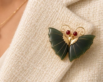 May include: A gold butterfly brooch with dark green wings and red detailing is pinned to a cream blazer. A gold necklace with a pearl pendant is also visible. The brooch is a decorative accessory.