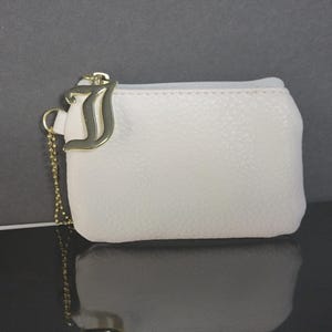 May include: A small, white coin purse with a textured surface. It features a gold-colored metal charm in the shape of the letter "J" and a delicate chain. The purse has a zipper closure.