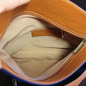 K&ouml;nnte beinhalten: Offene Ansicht einer braunen Lederhandtasche mit blauem Besatz. Der Innenraum ist beige mit einem kleinen braunen Lederetikett. Ein silberner Rei&szlig;verschluss verl&auml;uft entlang der Oberseite. Der Riemen ist braun und tan.