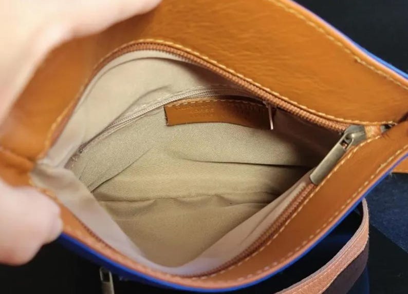 K&ouml;nnte beinhalten: Offene Ansicht einer braunen Lederhandtasche mit blauem Besatz. Der Innenraum ist beige mit einem kleinen braunen Lederetikett. Ein silberner Rei&szlig;verschluss verl&auml;uft entlang der Oberseite. Der Riemen ist braun und tan.