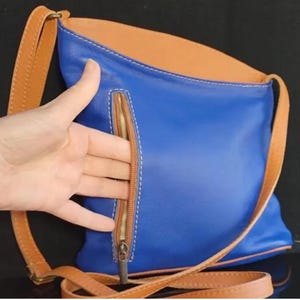 K&ouml;nnte beinhalten: Blaue Leder-Umh&auml;ngetasche mit beigem Innenraum und Riemen. Die Tasche hat eine beige Rei&szlig;verschlusstasche auf der Vorderseite und wei&szlig;e N&auml;hte. Der Riemen ist verstellbar und aus beigem Leder.