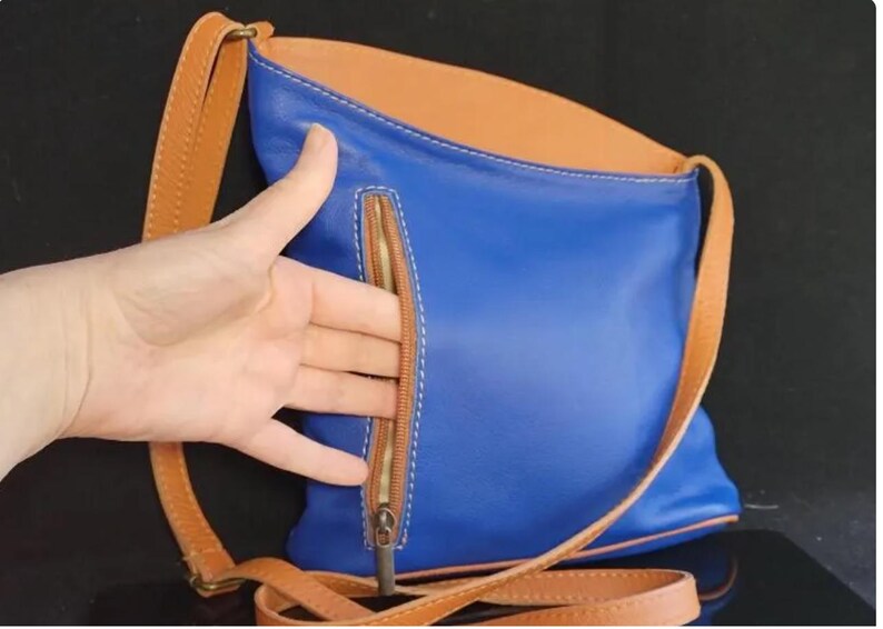 K&ouml;nnte beinhalten: Blaue Leder-Umh&auml;ngetasche mit beigem Innenraum und Riemen. Die Tasche hat eine beige Rei&szlig;verschlusstasche auf der Vorderseite und wei&szlig;e N&auml;hte. Der Riemen ist verstellbar und aus beigem Leder.