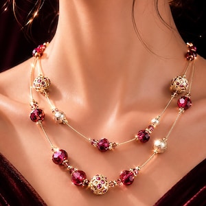 May include: A double-strand gold-tone necklace featuring faceted deep red crystal beads and ornate gold-tone filigree spheres. The necklace is designed with alternating red and gold elements along thin gold-tone chains, creating a layered look.