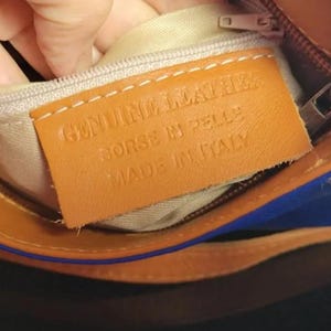K&ouml;nnte beinhalten: Nahaufnahme des Innenraums einer braunen Ledertasche mit beigem Futter. Ein rechteckiges Lederetikett lautet "GENUINE LEATHER BORSE IN PELLE MADE IN ITALY". Die Tasche hat einen Rei&szlig;verschluss und blaue Akzente.