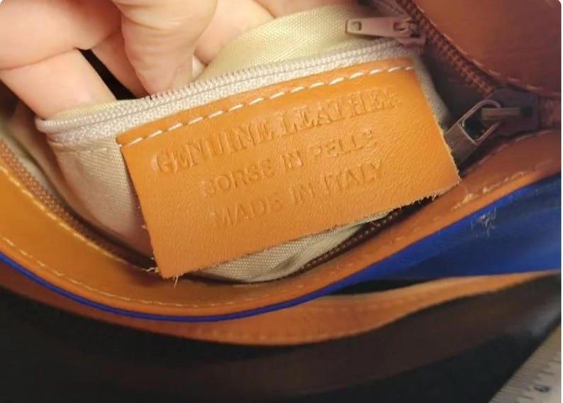 K&ouml;nnte beinhalten: Nahaufnahme des Innenraums einer braunen Ledertasche mit beigem Futter. Ein rechteckiges Lederetikett lautet "GENUINE LEATHER BORSE IN PELLE MADE IN ITALY". Die Tasche hat einen Rei&szlig;verschluss und blaue Akzente.