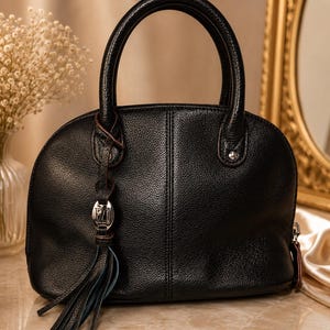 May include: Black leather handbag with a rounded shape and two top handles. The bag features a decorative tassel with a silver-tone charm and a vertical seam down the center. The bag is sitting on a marble surface.