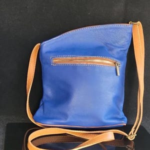 K&ouml;nnte beinhalten: Eine blaue Leder-Umh&auml;ngetasche mit einem beigefarbenen Riemen und Rei&szlig;verschluss. Die Tasche hat eine rechteckige Form mit abgerundeten Kanten. Der Rei&szlig;verschluss ist beige und die N&auml;hte sind wei&szlig;. Die Tasche ist aus glattem Leder.
