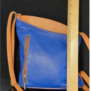 K&ouml;nnte beinhalten: Blaue Leder-Umh&auml;ngetasche mit braunem Innenraum und Riemen. Die Tasche hat eine Rei&szlig;verschlusstasche auf der Vorderseite und ist ca. 25 cm hoch.