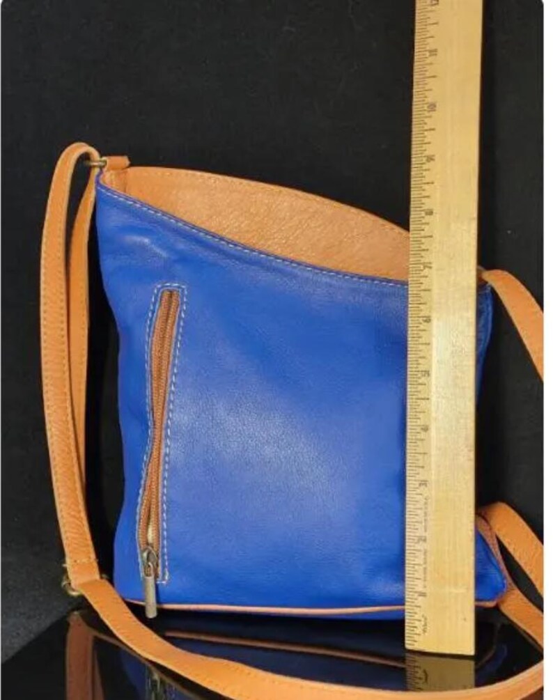 K&ouml;nnte beinhalten: Blaue Leder-Umh&auml;ngetasche mit braunem Innenraum und Riemen. Die Tasche hat eine Rei&szlig;verschlusstasche auf der Vorderseite und ist ca. 25 cm hoch.