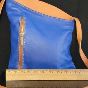 K&ouml;nnte beinhalten: Blaue Leder-Umh&auml;ngetasche mit beigem Innenraum und Riemen. Die Tasche hat eine Rei&szlig;verschlusstasche vorne mit braunem Rei&szlig;verschluss und wei&szlig;en N&auml;hten. Ein Holzlineal befindet sich am unteren Bildrand.