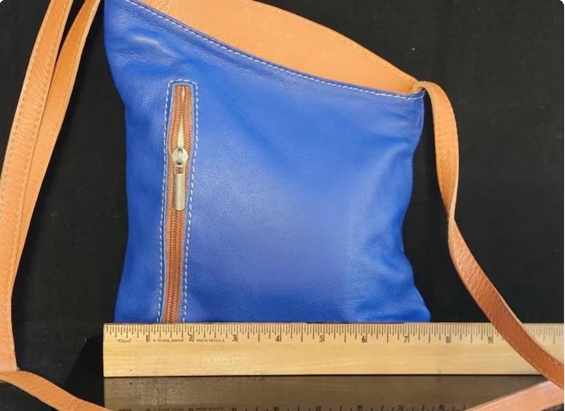 K&ouml;nnte beinhalten: Blaue Leder-Umh&auml;ngetasche mit beigem Innenraum und Riemen. Die Tasche hat eine Rei&szlig;verschlusstasche vorne mit braunem Rei&szlig;verschluss und wei&szlig;en N&auml;hten. Ein Holzlineal befindet sich am unteren Bildrand.