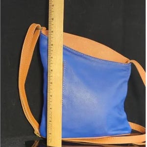 K&ouml;nnte beinhalten: Eine blaue und beige Crossbody-Tasche aus Leder mit einem beigefarbenen Riemen. Die Tasche ist etwa 25 cm hoch. Sie hat eine rechteckige Form mit gew&ouml;lbtem Oberteil. Der Riemen ist d&uuml;nn und verstellbar.