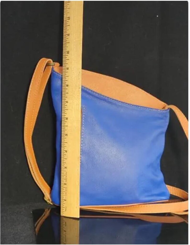 K&ouml;nnte beinhalten: Eine blaue und beige Crossbody-Tasche aus Leder mit einem beigefarbenen Riemen. Die Tasche ist etwa 25 cm hoch. Sie hat eine rechteckige Form mit gew&ouml;lbtem Oberteil. Der Riemen ist d&uuml;nn und verstellbar.