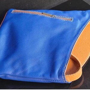 K&ouml;nnte beinhalten: Eine blaue Leder-Clutch mit orangefarbenem Innenraum und Riemen. Die Clutch hat einen Rei&szlig;verschluss mit braunem Rei&szlig;verschluss und wei&szlig;en N&auml;hten. Die Kanten sind mit braunen N&auml;hten versehen.