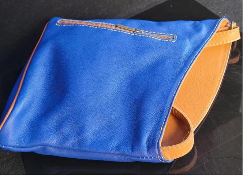 K&ouml;nnte beinhalten: Eine blaue Leder-Clutch mit orangefarbenem Innenraum und Riemen. Die Clutch hat einen Rei&szlig;verschluss mit braunem Rei&szlig;verschluss und wei&szlig;en N&auml;hten. Die Kanten sind mit braunen N&auml;hten versehen.