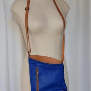 K&ouml;nnte beinhalten: Eine blaue Leder-Umh&auml;ngetasche mit einem braunen Lederriemen und Rei&szlig;verschluss. Die Tasche wird auf einer Schaufensterpuppe pr&auml;sentiert. Die Innenseite der Tasche ist braun.