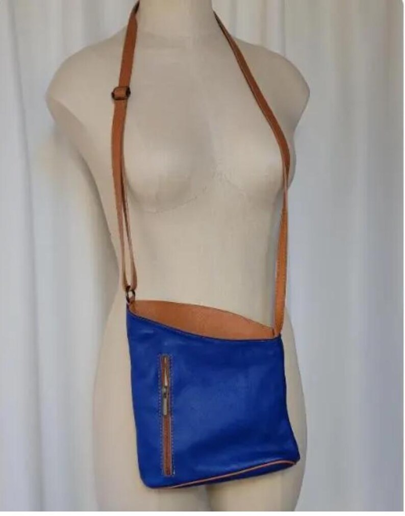 K&ouml;nnte beinhalten: Eine blaue Leder-Umh&auml;ngetasche mit einem braunen Lederriemen und Rei&szlig;verschluss. Die Tasche wird auf einer Schaufensterpuppe pr&auml;sentiert. Die Innenseite der Tasche ist braun.