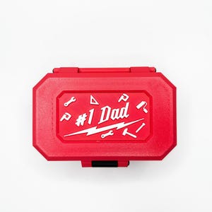 Könnte beinhalten: Eine rote Werkzeugkiste mit weißer Aufschrift "#1 Dad" und verschiedenen Werkzeugsymbolen. Die Werkzeugkiste ist rechteckig mit einem Klappdeckel und einem schwarzen Verschluss. Der Hintergrund ist weiß.
