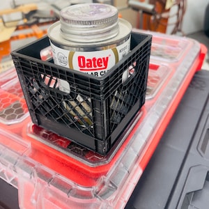 Puede incluir: Una caja de pl&aacute;stico negro que contiene una lata de cemento PVC transparente Oatey. La lata tiene una tapa plateada y una etiqueta blanca y dorada. La caja est&aacute; colocada sobre un recipiente de pl&aacute;stico rojo y transparente.