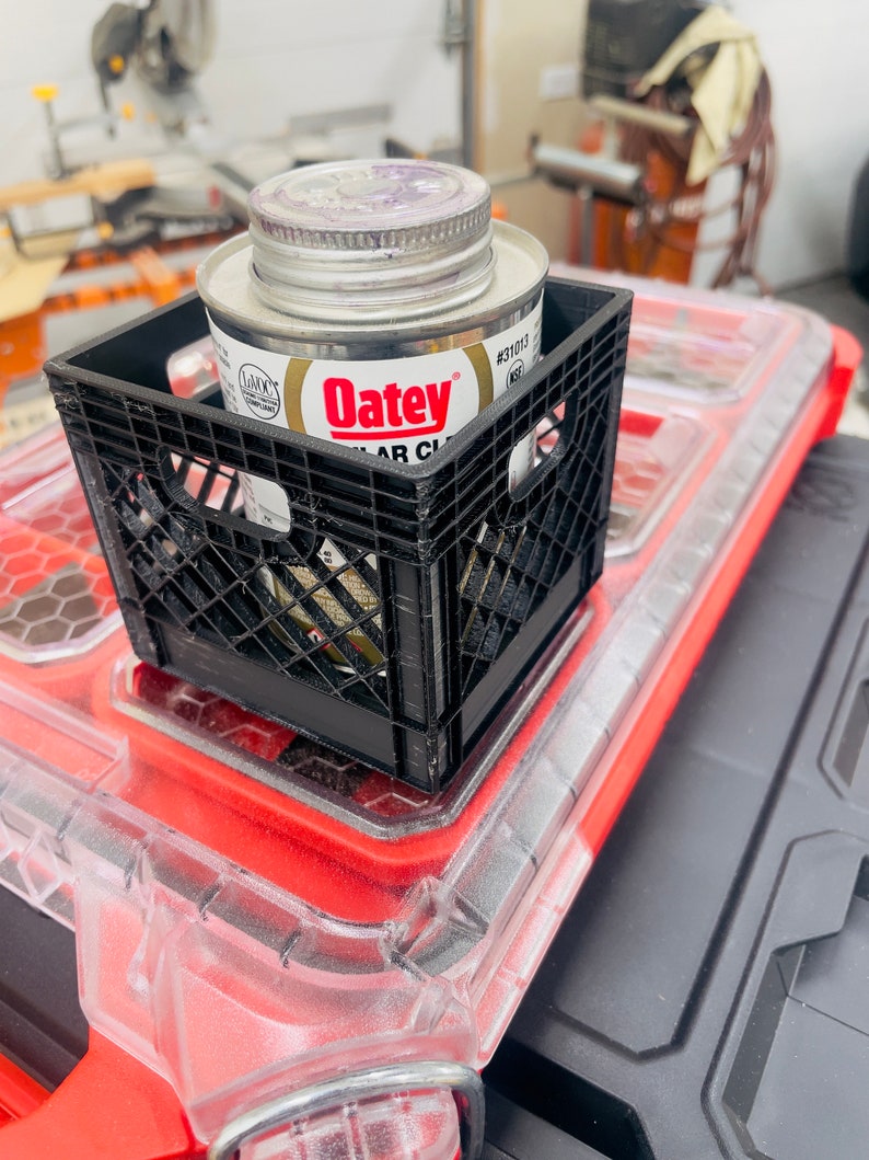 Puede incluir: Una caja de pl&aacute;stico negro que contiene una lata de cemento PVC transparente Oatey. La lata tiene una tapa plateada y una etiqueta blanca y dorada. La caja est&aacute; colocada sobre un recipiente de pl&aacute;stico rojo y transparente.