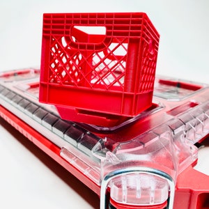 Puede incluir: Una peque&ntilde;a caja de leche de pl&aacute;stico roja se encuentra encima de un objeto de pl&aacute;stico rojo y transparente. La caja es una caja abierta con un patr&oacute;n de rejilla. El objeto de abajo tiene una parte superior de pl&aacute;stico transparente y detalles rojos.