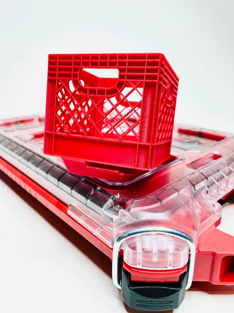 Puede incluir: Una peque&ntilde;a caja de leche de pl&aacute;stico roja se encuentra encima de un objeto de pl&aacute;stico rojo y transparente. La caja es una caja abierta con un patr&oacute;n de rejilla. El objeto de abajo tiene una parte superior de pl&aacute;stico transparente y detalles rojos.
