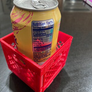Puede incluir: Una lata amarilla con tapa plateada se encuentra dentro de una peque&ntilde;a caja de pl&aacute;stico roja. La lata tiene una etiqueta de informaci&oacute;n nutricional visible. La caja es cuadrada con un dise&ntilde;o de celos&iacute;a.