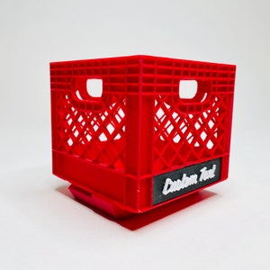 Puede incluir: Una caja de pl&aacute;stico roja con forma rectangular y dise&ntilde;o de celos&iacute;a. La caja tiene dos grandes aberturas para las asas en los laterales. Una etiqueta negra con la inscripci&oacute;n "Custom Text" en blanco est&aacute; adherida en la parte delantera.