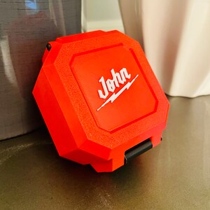 May include: A bright orange, rectangular-shaped storage case with a hinged lid. The case has a white inscription that reads "John" with a lightning bolt graphic.