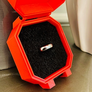 May include: A red ring box with a black velvet interior. A silver ring is displayed inside the box.