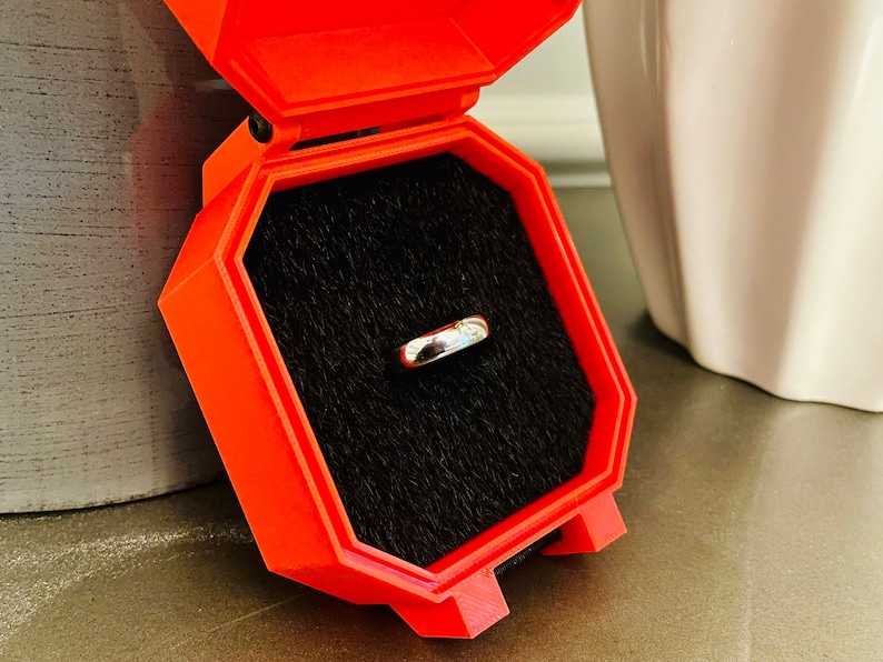 May include: A red ring box with a black velvet interior. A silver ring is displayed inside the box.