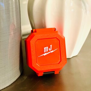 May include: A bright orange, octagonal-shaped case with a black latch. The case has the letters "MJ" and a lightning bolt logo in white.
