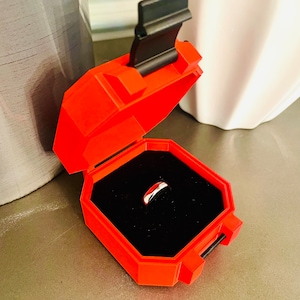 May include: A red ring box with a black lid and a silver ring inside. The box is open and the ring is visible in the center of the box.