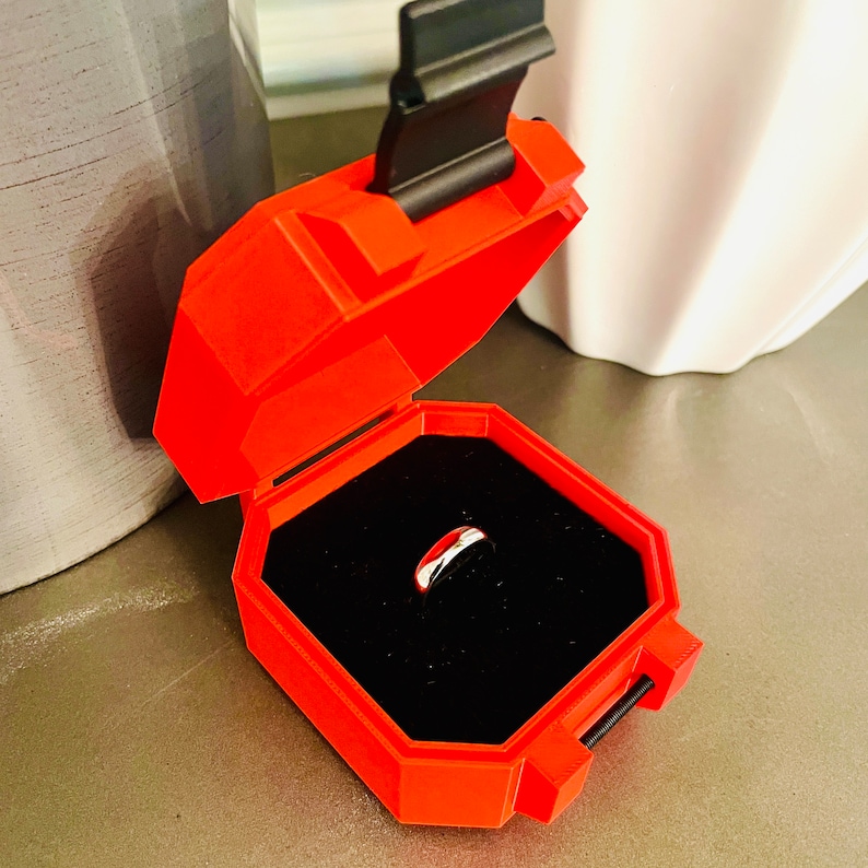 May include: A red ring box with a black lid and a silver ring inside. The box is open and the ring is visible in the center of the box.