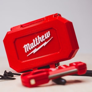May include: A red toolbox with the word "Matthew" printed in white on the top. The toolbox is sitting on a wooden surface with some screws and a red tool.