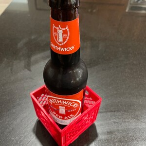 Puede incluir: Una botella de Smithwick's Red Ale en una peque&ntilde;a caja de pl&aacute;stico roja. La botella tiene una tapa plateada y una etiqueta roja con texto blanco y un escudo. La caja es cuadrada y tiene un patr&oacute;n de rejilla.