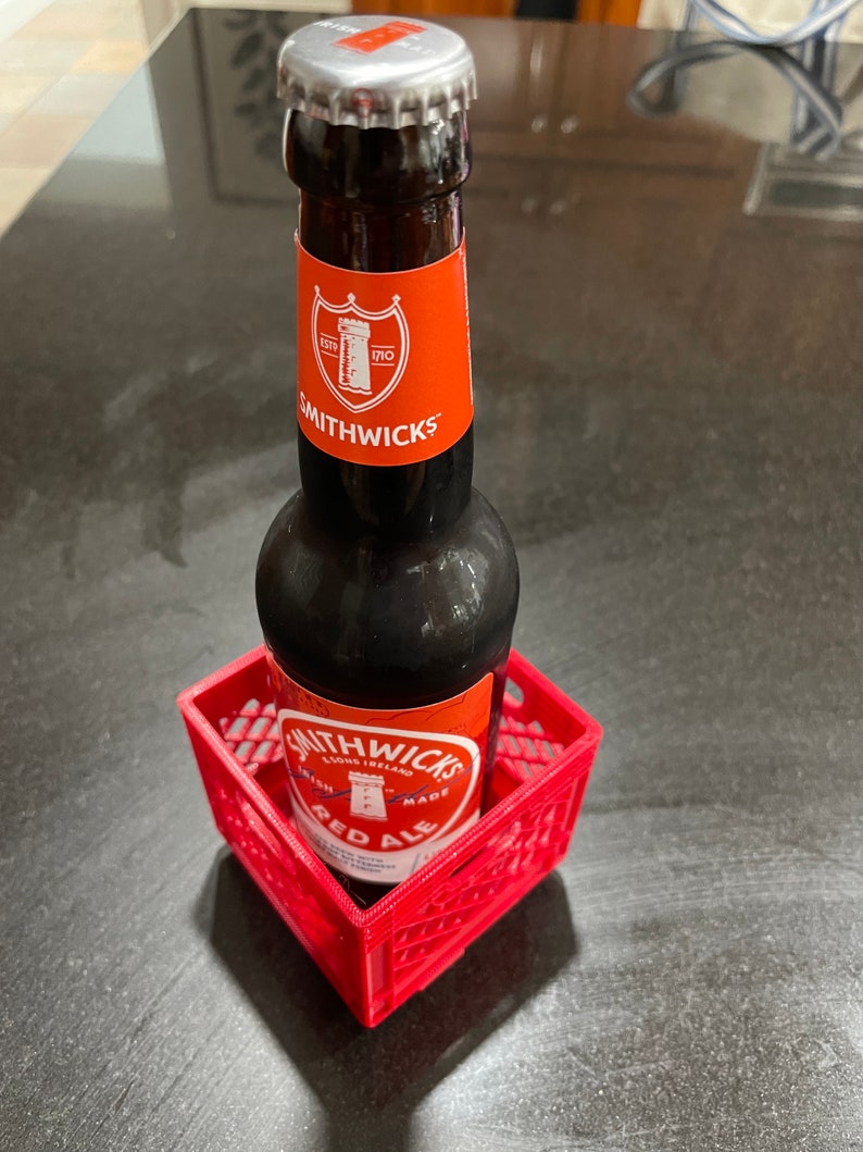 Puede incluir: Una botella de Smithwick's Red Ale en una peque&ntilde;a caja de pl&aacute;stico roja. La botella tiene una tapa plateada y una etiqueta roja con texto blanco y un escudo. La caja es cuadrada y tiene un patr&oacute;n de rejilla.