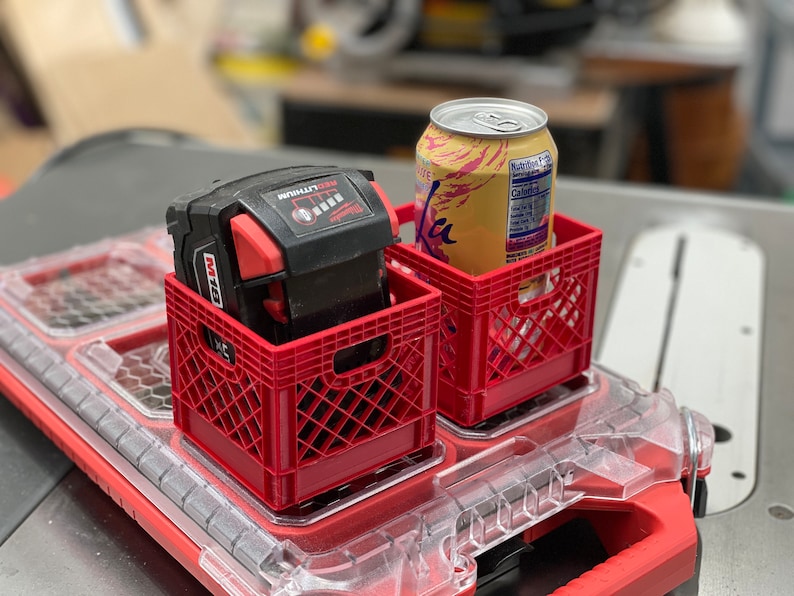 Puede incluir: Dos peque&ntilde;as cajas de pl&aacute;stico rojas, una con una bater&iacute;a de herramienta el&eacute;ctrica negra y roja, la otra con una lata de agua con gas La Croix. Las cajas est&aacute;n sobre una caja de herramientas de pl&aacute;stico roja y transparente, con una sierra de mesa al fondo.