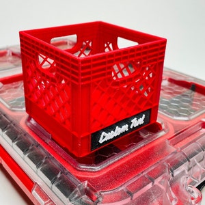 Puede incluir: Una peque&ntilde;a caja de pl&aacute;stico roja con un dise&ntilde;o de celos&iacute;a, colocada sobre una superficie transparente te&ntilde;ida de rojo. La caja tiene una etiqueta negra con la inscripci&oacute;n "Custom Text" en blanco. Es una versi&oacute;n en miniatura de una caja de leche.