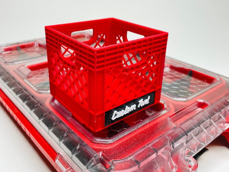 Puede incluir: Una peque&ntilde;a caja de pl&aacute;stico roja con un dise&ntilde;o de celos&iacute;a, colocada sobre una superficie transparente te&ntilde;ida de rojo. La caja tiene una etiqueta negra con la inscripci&oacute;n "Custom Text" en blanco. Es una versi&oacute;n en miniatura de una caja de leche.