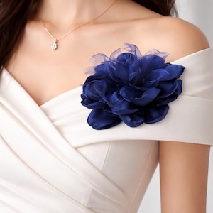 以下が含まれることがあります： 白のドレスにネイビーブルーの花のアクセントのクローズアップ。花は重ねられた生地で作られており、ドレスの肩に付けられています。小さなペンダントが付いたゴールドのネックレスも表示されています。