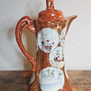 Puede incluir: Una tetera de porcelana japonesa vintage con un esmalte marrón y dorado. La tetera presenta un diseño floral y una escena de un puente y edificios. La tetera tiene una tapa con un pomo.