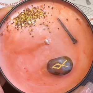 Op de afbeelding: Roze kaars met een metalen deksel. De kaars heeft een bruine spijker, een kleine bruine steen met een gouden rune en gedroogde kruiden erop.