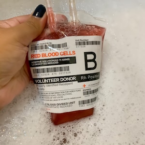 Puede incluir: Una bolsa de sangre roja con una etiqueta blanca que dice "RED BLOOD CELLS" y "VOLUNTEER DONOR". La bolsa está etiquetada con una fecha de recolección, un número de unidad y un tipo de sangre. La bolsa está en un fregadero lleno de agua jabonosa.