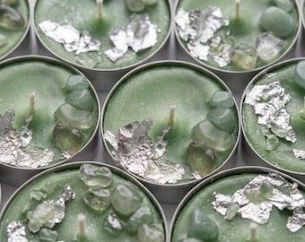 May include: A collection of round tea light candles in metal cups, each featuring light green wax, a central white wick, small polished green stones, and scattered pieces of metallic silver leaf foil embedded on the top surface of the wax.