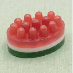 May include: An oval-shaped massage soap bar featuring a translucent red top layer with twelve rounded protrusions, a solid white middle layer, and a translucent green bottom layer, designed with a layered color scheme resembling a watermelon slice.