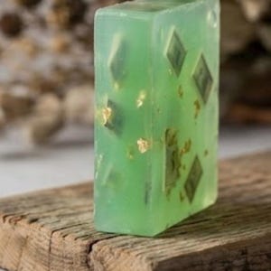 May include: A rectangular bar of translucent light green soap featuring embedded miniature green paper currency replicas and small flecks of gold leaf throughout the semi-transparent body of the cleansing bar.