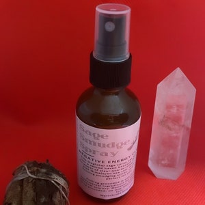 May include: A brown glass bottle with a black spray top and a white label that reads "Sage Smudge Spray" and "NEGATIVE ENERGY CLEANSING". The bottle is sitting on a red surface next to a clear quartz crystal.
