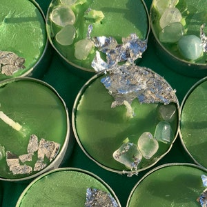 May include: Close-up of several green tea light candles in metal holders. Each candle is decorated with clear and green stones and silver foil. The candles are arranged on a green surface.