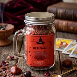May include: A clear glass mason jar with a handle filled with coarse red salt. The jar features a rectangular orange label with a black witch hat icon and text that reads Red Salt, Passion Self Love Root Chakra, and Waterbruja Etsy Com.